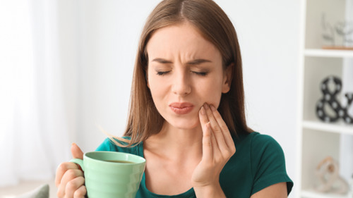 Young woman holding a coffee mug while wincing and touching her jaw in tooth pain